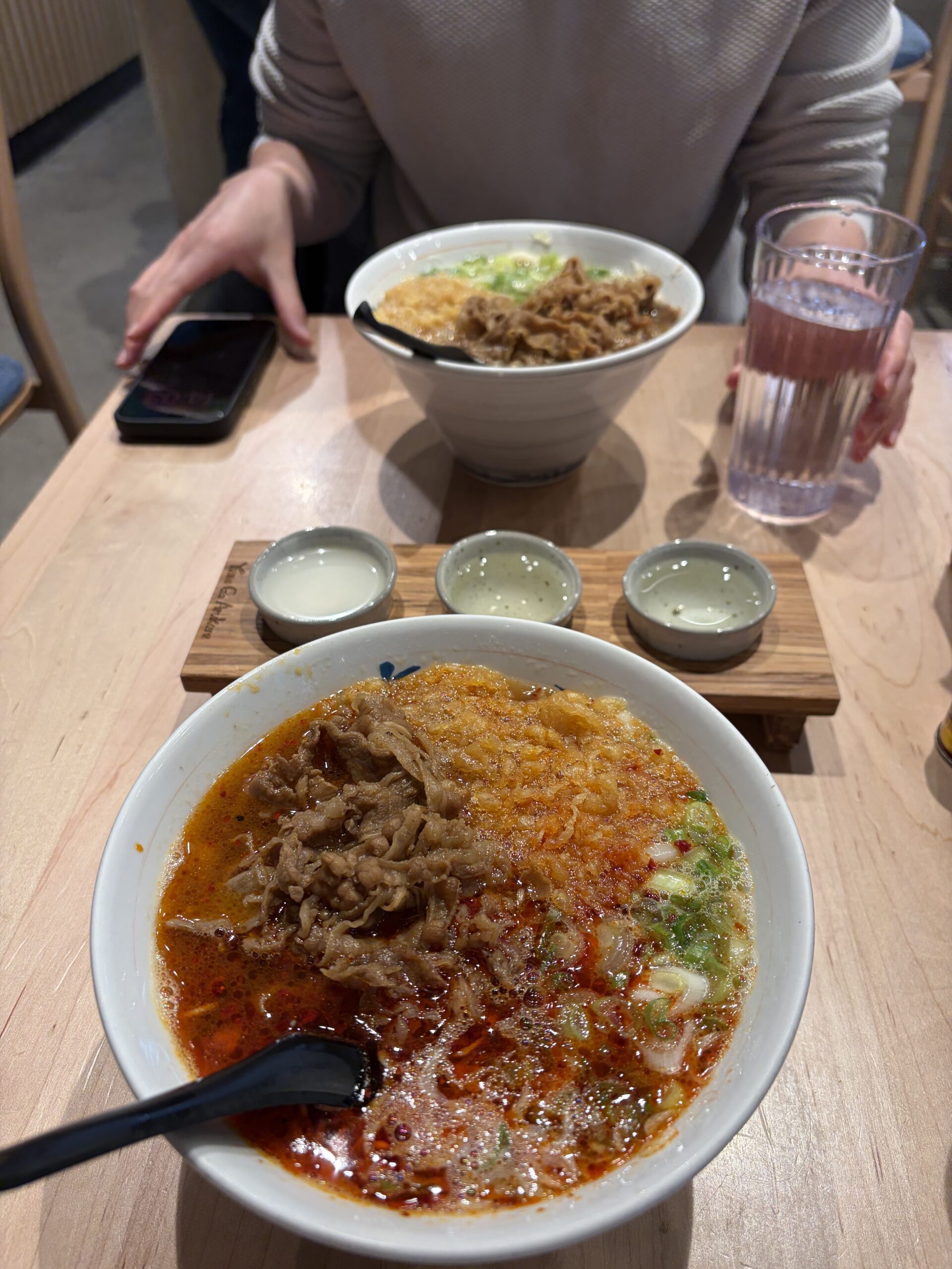 Two bowls of Yume Ga Arukara niku udon—one regular and one spicy hot—separated by a flight of sake glasses on a wooden table.