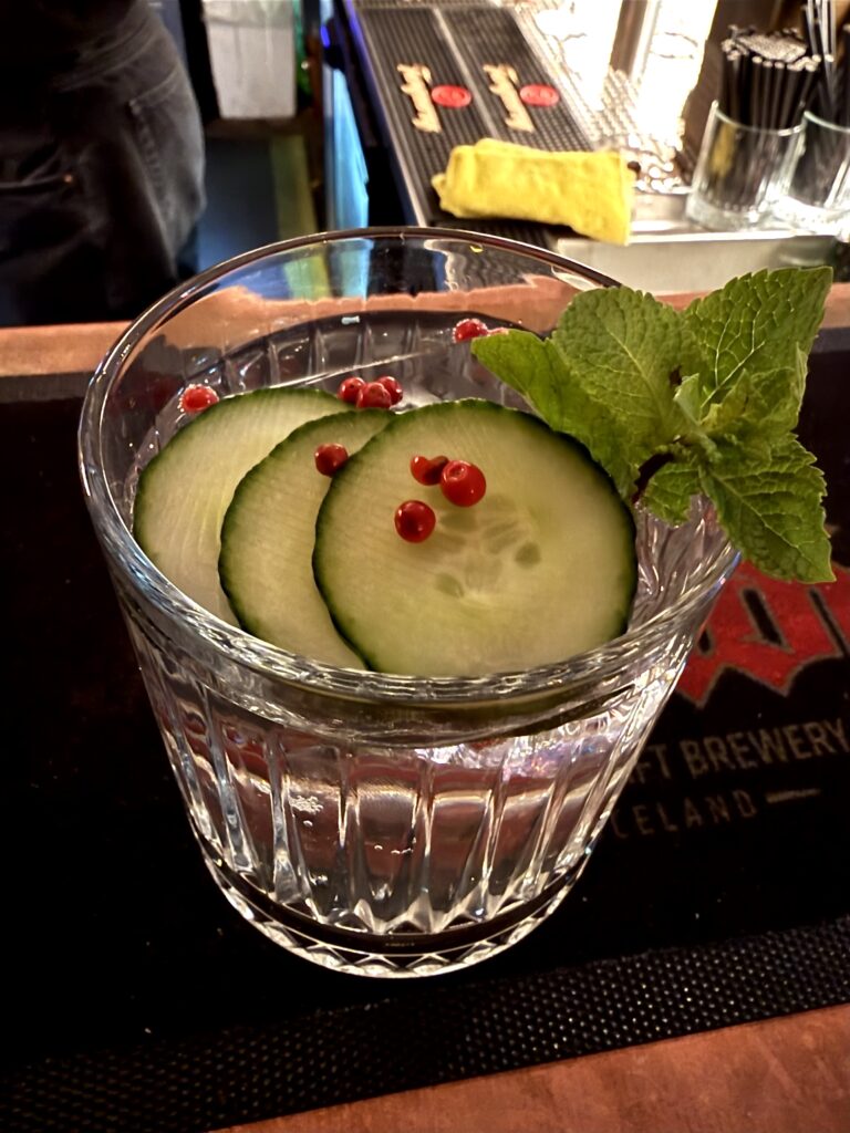 A clear cocktail in a clear glass topped with cucumbers, berries, and mint. 