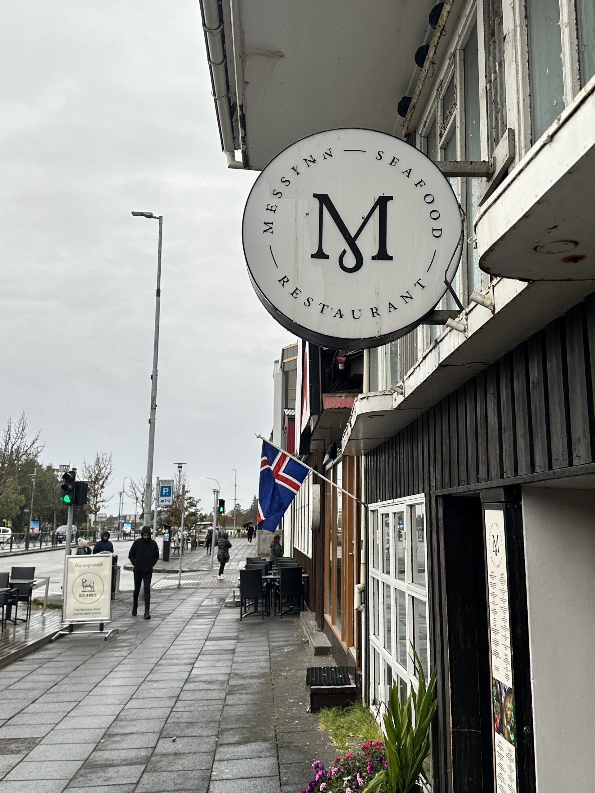 The exterior of Messinn Seafood Restaurant in Reykjavik, showing the circular storefront sign with a large "M" logo hanging over a damp sidewalk on a cloudy day.