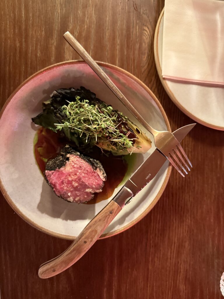 A plated dish on a wooden table featuring a thick, medium-rare cut of Icelandic lamb with a dark crust, served alongside grilled cabbage topped with microgreens and a rich jus.