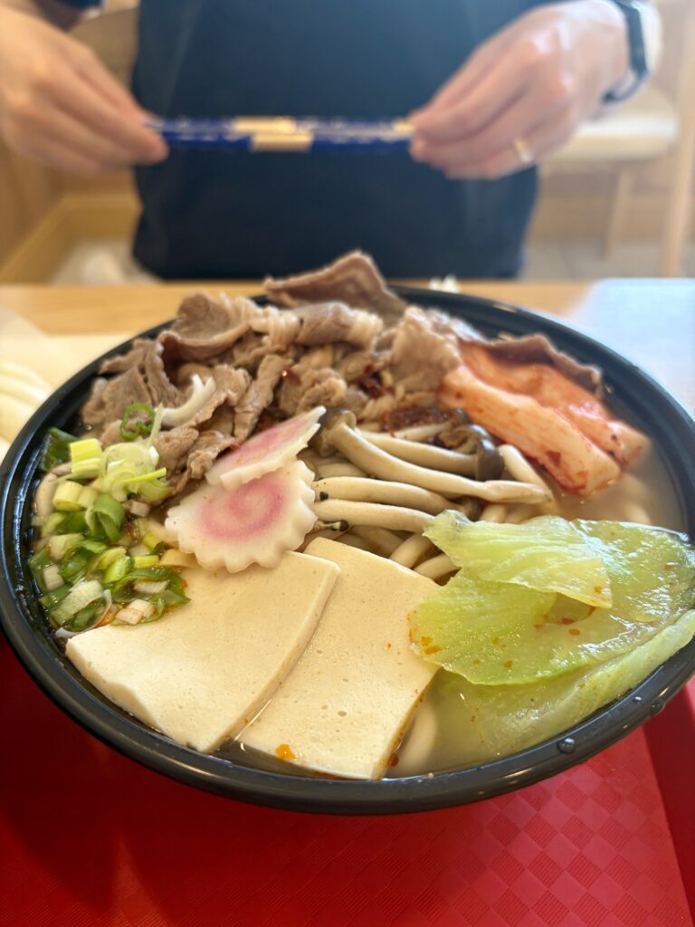Beef noodle soup in a black plastic bowl topped with thinly sliced beef, mushrooms, naruto fish cake, tofu, green onions, and imitation crab, 