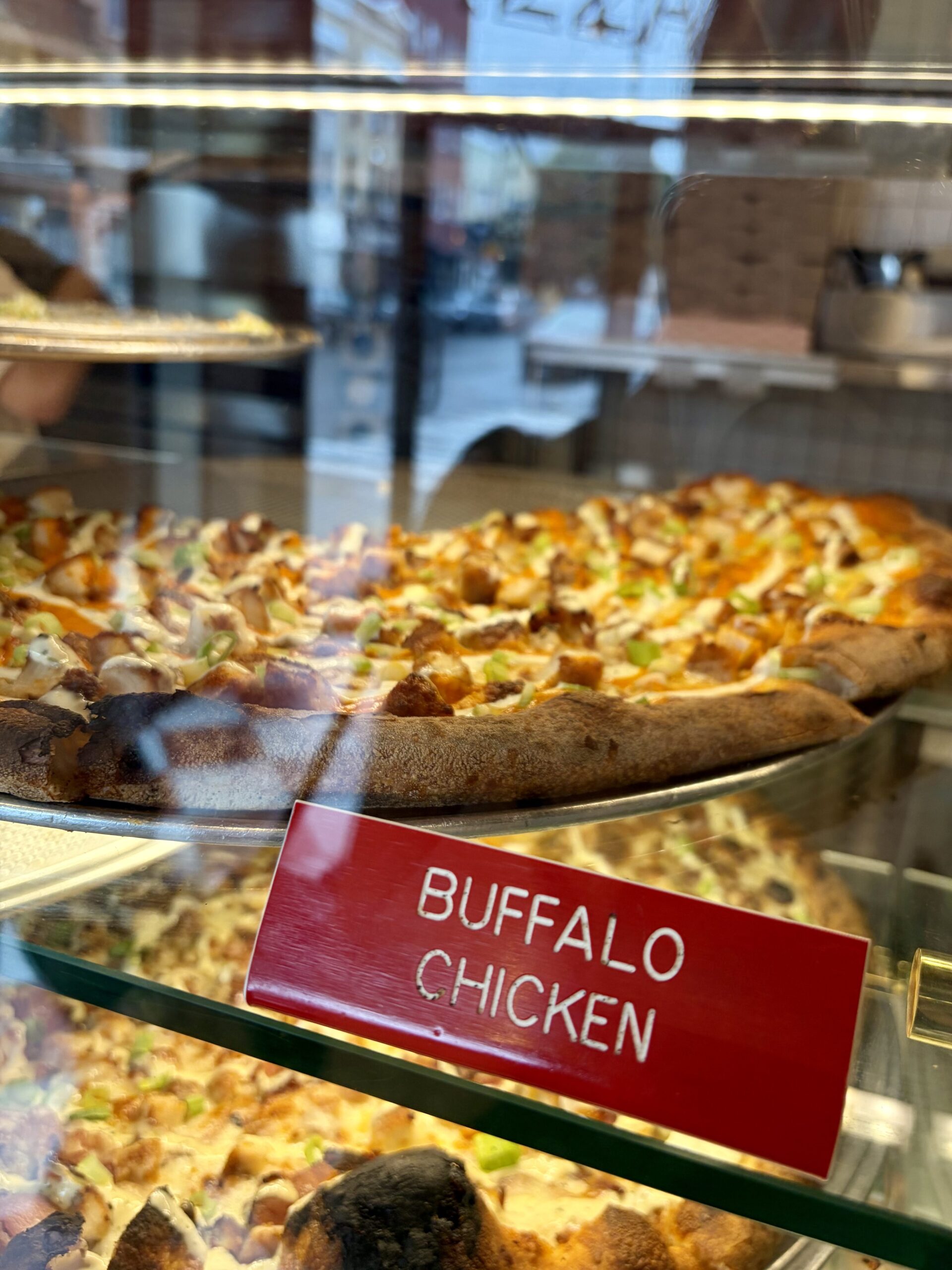 Buffalo Chicken pizza in a display case with a red and white sign. 
