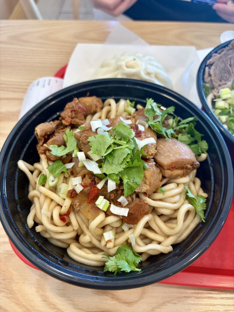 Noodles in a black plastic bowl topped with chicken highs, peppers,  cilantro, and green onion. 