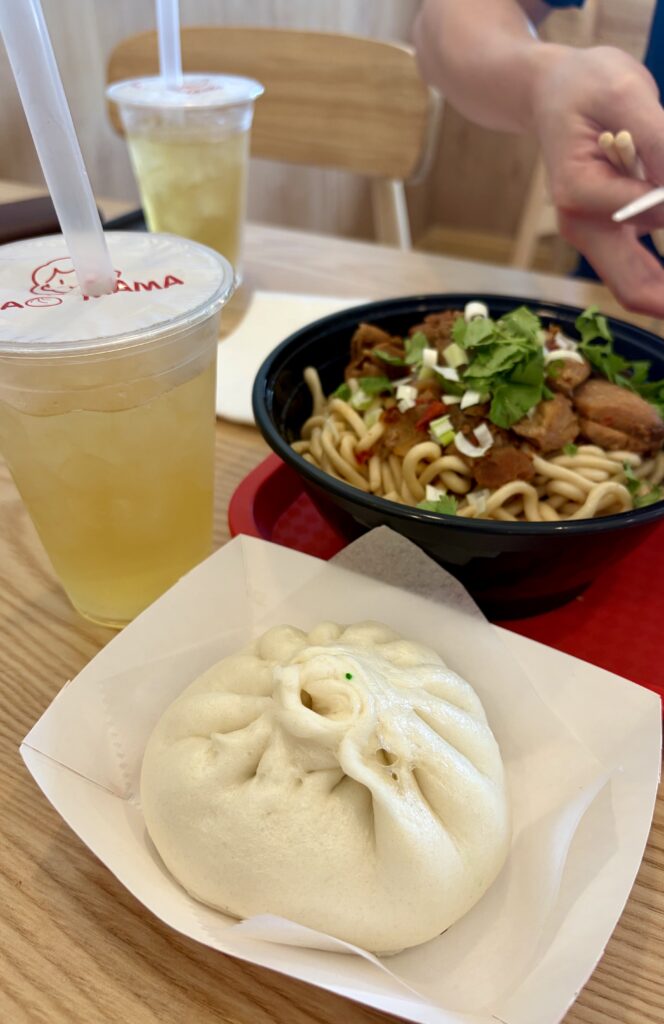 Picture of the lunch special.Ice tea with the noodle bowl next to a large white bao bun,. 